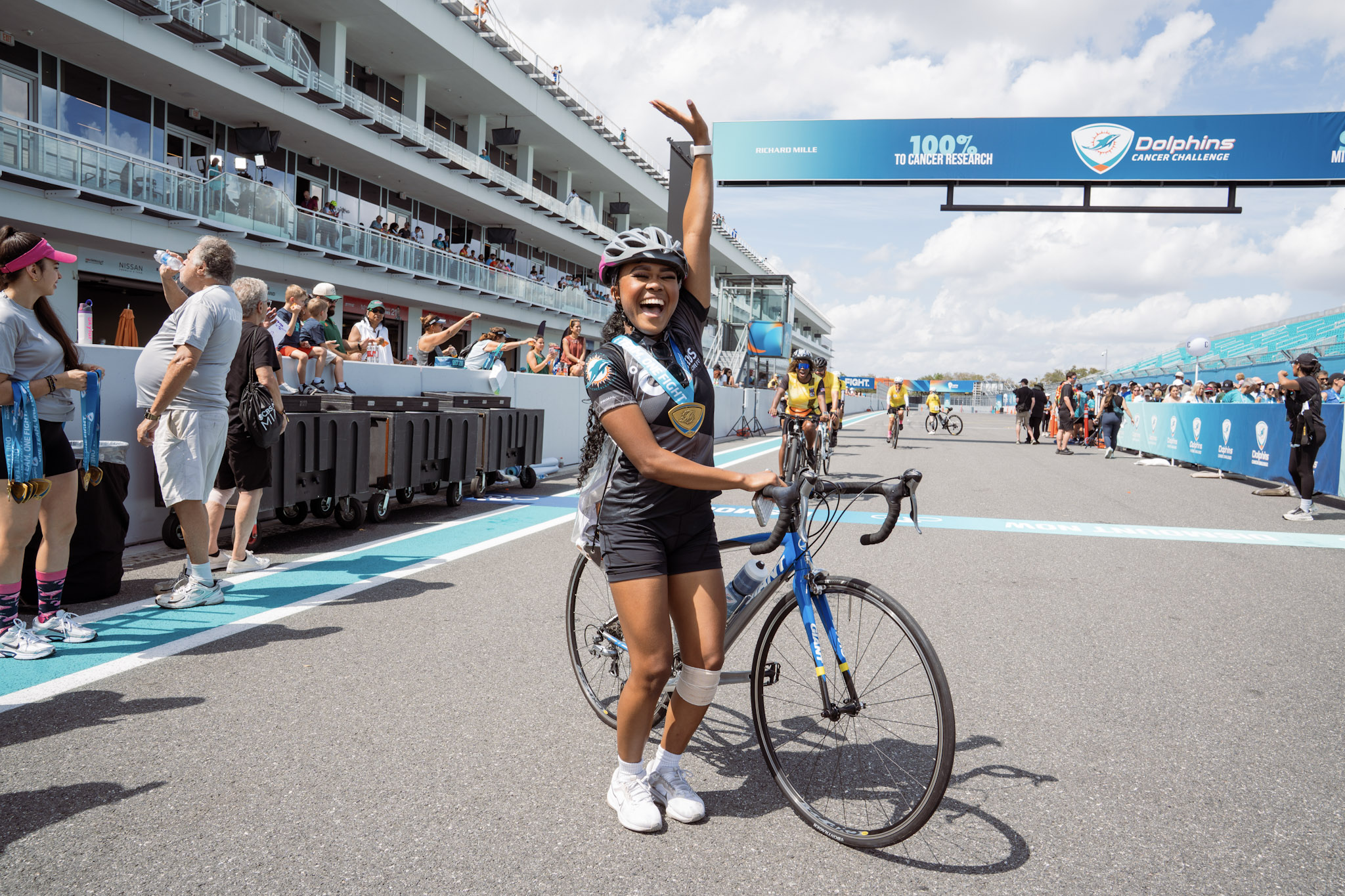 A female bicyclist at the finish line at Dolphins Cancer Challenge, leaning on her bike and raising her left arm in triump